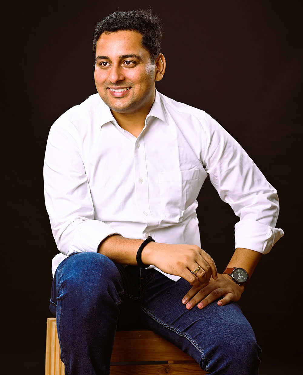 Coach Rajendra smiling in a white shirt and blue jeans, sitting on wooden stool, exuding confidence and approachability.